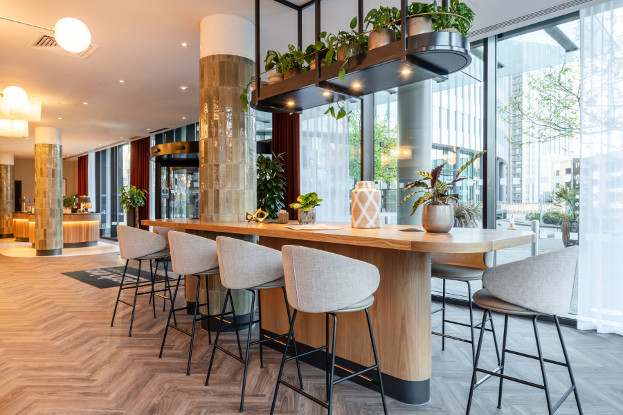 Modern hotel lobby or cafe with a long wooden bar-height table, beige stools, and potted plants near floor-to-ceiling windows bathed in natural light.