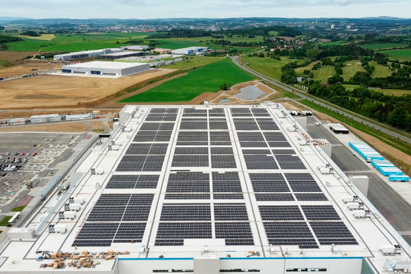 Aerial view of a large industrial roof covered with solar panels, surrounded by green fields and roads around.