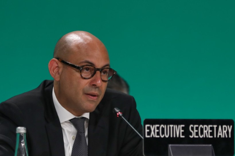 Bald man in a suit and glasses speaking into a microphone at a panel, with an 'Executive Secretary' sign nearby.
