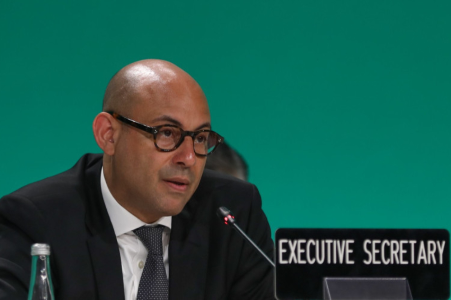 Bald man in a suit and glasses speaking into a microphone at a panel, with an 'Executive Secretary' sign nearby.