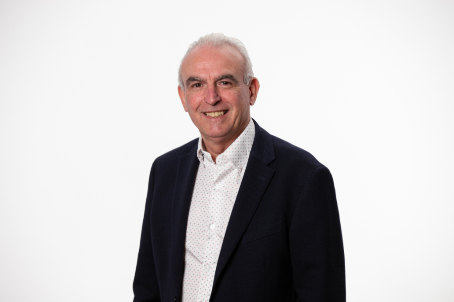 Friendly head-and-shoulders portrait of a smiling older man in a navy blazer and white patterned shirt against a white background, professional headshot.