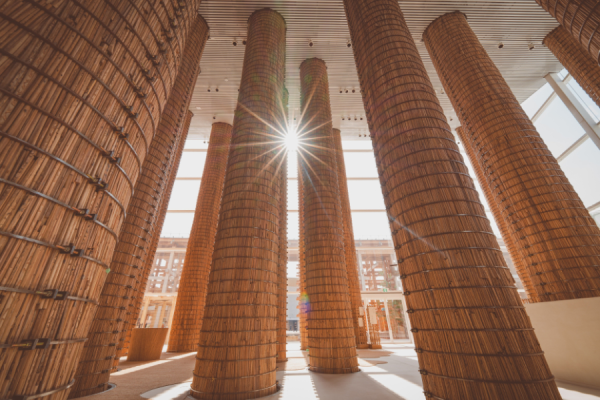 Sunlit atrium with tall cylindrical woven straw columns surrounding a central open space.