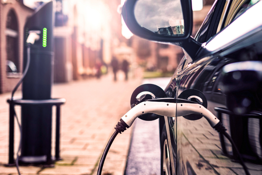 Electric vehicle charging on a city street; close-up of the charging plug connected to the car side panel, with a blurred urban background.