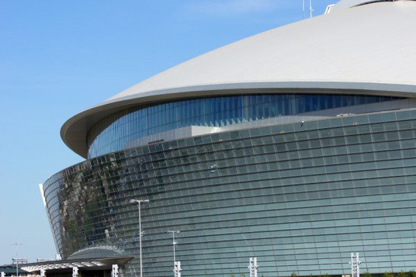 Curved glass-and-metal facade of a large modern museum under a pale domed roof, set against a blue sky.