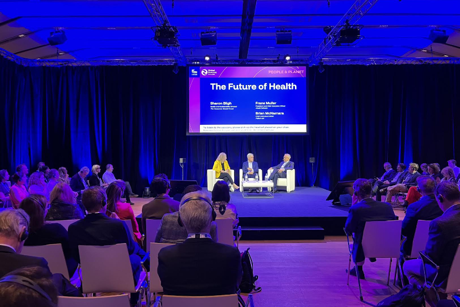 Panel discussion on a blue-lit stage with three speakers and an audience watching; screen shows 'The Future of Health'.