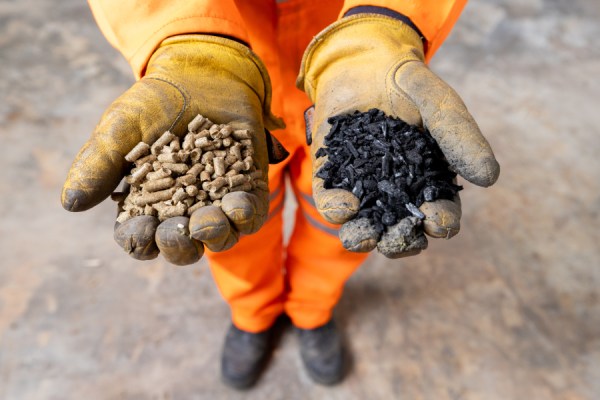 Worker in orange safety gear offers two handfuls of pellets: light brown wood pellets and dark black coal/charcoal pellets.