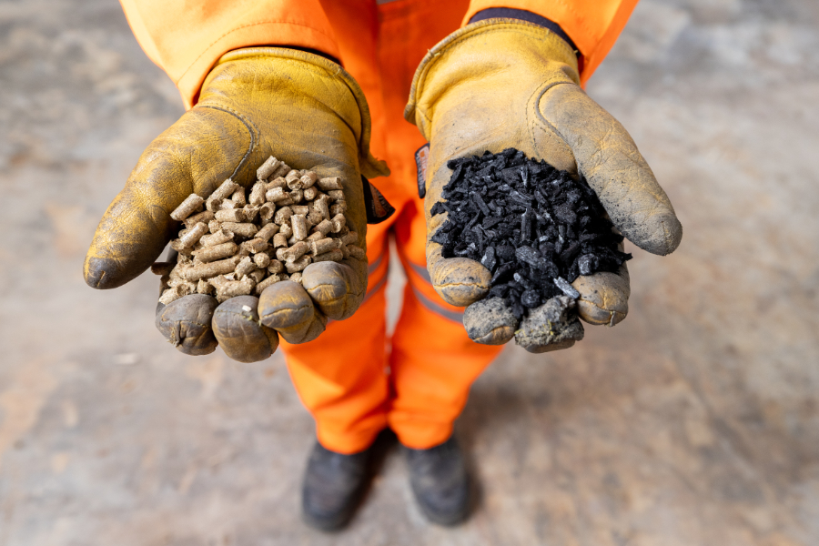 Worker in orange safety gear offers two handfuls of pellets: light brown wood pellets and dark black coal/charcoal pellets.