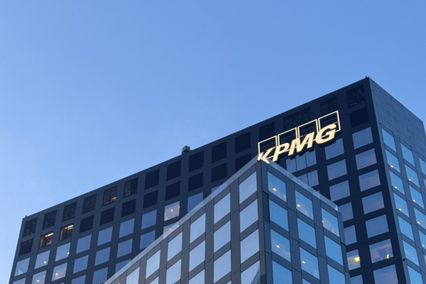 Tall glass office building with a lit KPMG logo on the roof against a clear blue sky and sunset reflections in the windows