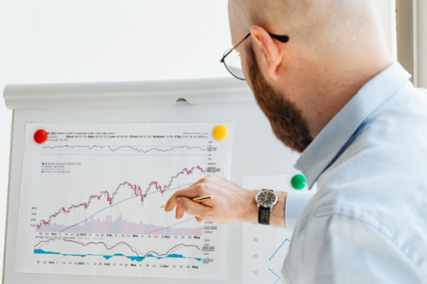 Man with glasses and beard analyzes a financial chart on a whiteboard, using a pencil to point at data.
