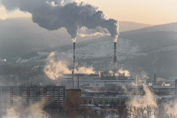 Factory complex with tall smokestacks emitting dense smoke over a city with snow-covered hills in the background.