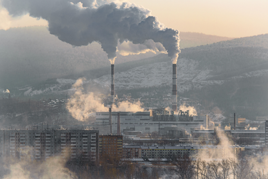 Factory complex with tall smokestacks emitting dense smoke over a city with snow-covered hills in the background.