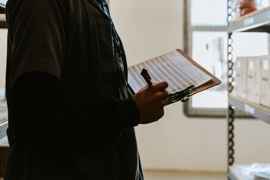 Person in dark clothing holds a clipboard and pen, inspecting inventory in a warehouse aisle.