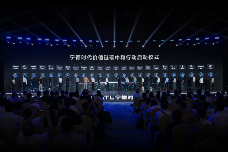 A large conference stage with dozens of people standing in a line on stage, illuminated by blue lighting, while a seated audience watches and Chinese text is displayed on a wide screen behind them.