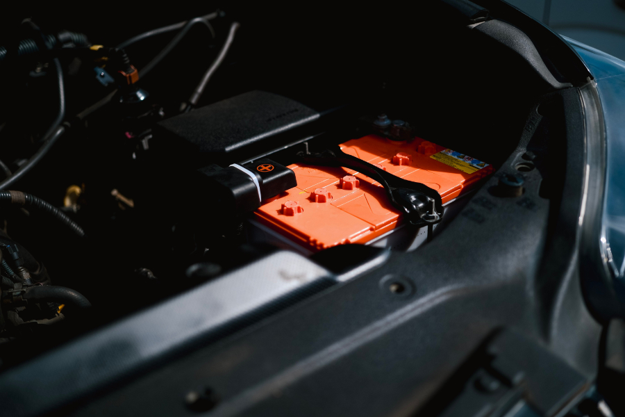 Car engine bay with a bright orange car battery visible under the open hood, cables and components surrounding it.